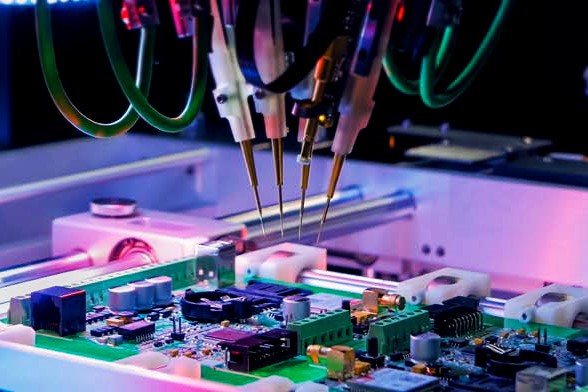 Close-up of a robotic machine with multiple nozzles assembling a power inductor on a green circuit board, under colorful lighting in an electronics manufacturing environment. by Standex Edge