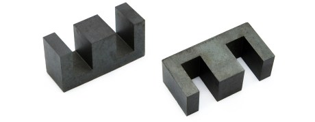 Two dark gray metal objects with three rectangular prongs each are positioned on a white background. Designed to maximize clearance and creepage, the left object stands upright while the right one lies flat, showing a similar shape from another angle. by Standex Edge