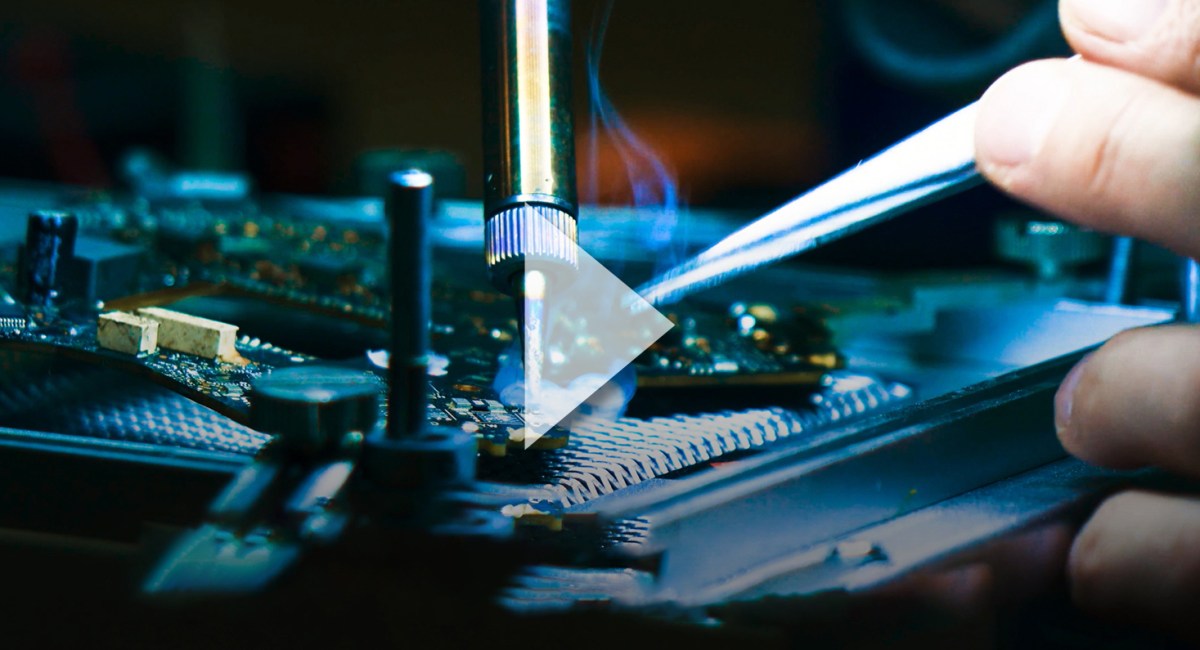 Close-up of hands using tweezers and a soldering iron to work on a circuit board, highlighting semiconductor tools, with a white play button overlay in the center, suggesting this is a video thumbnail. by Standex Edge